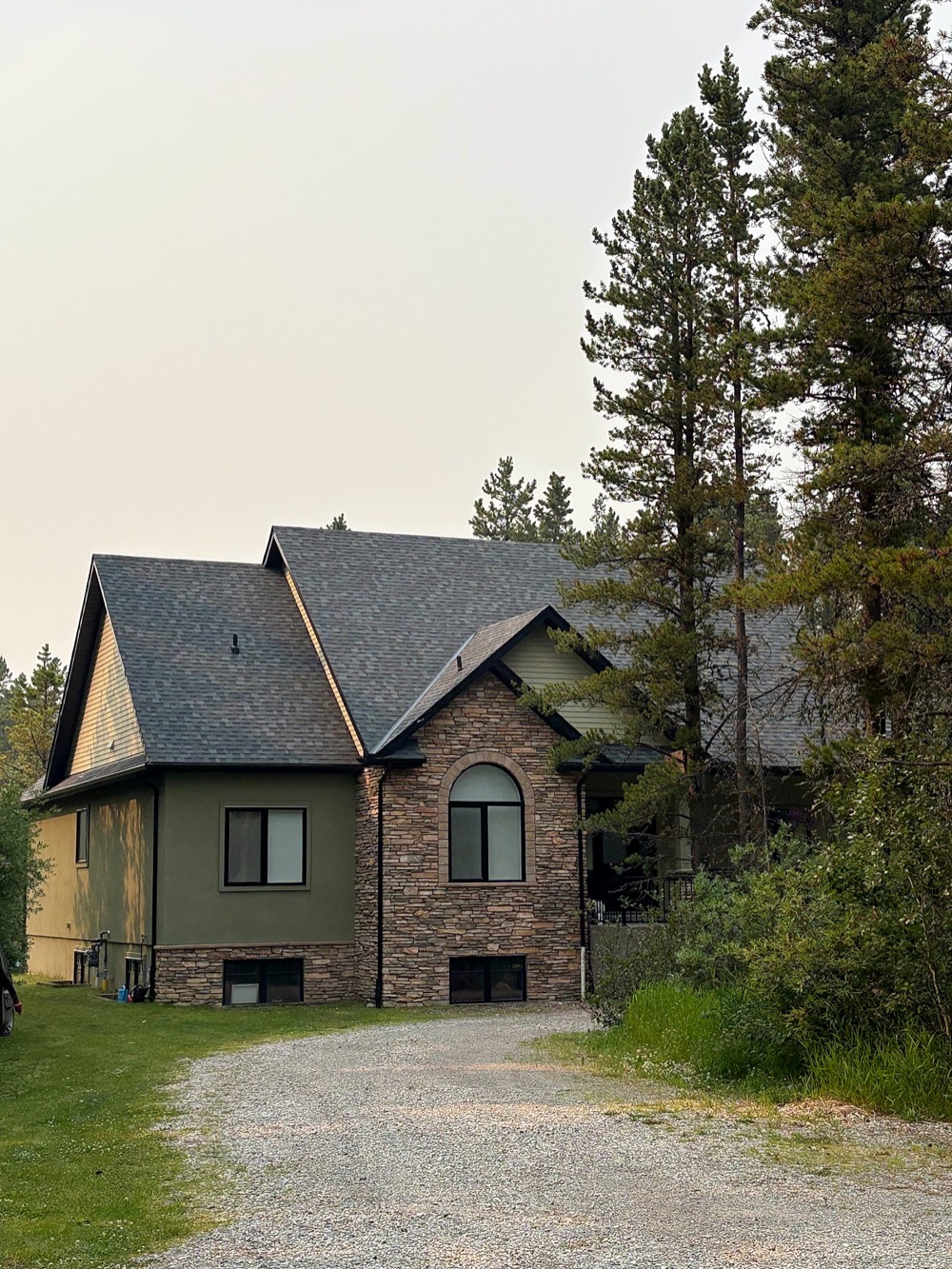 Two-storey stone and stucco house with a dark shingle roof, gravel drive and spruce trees