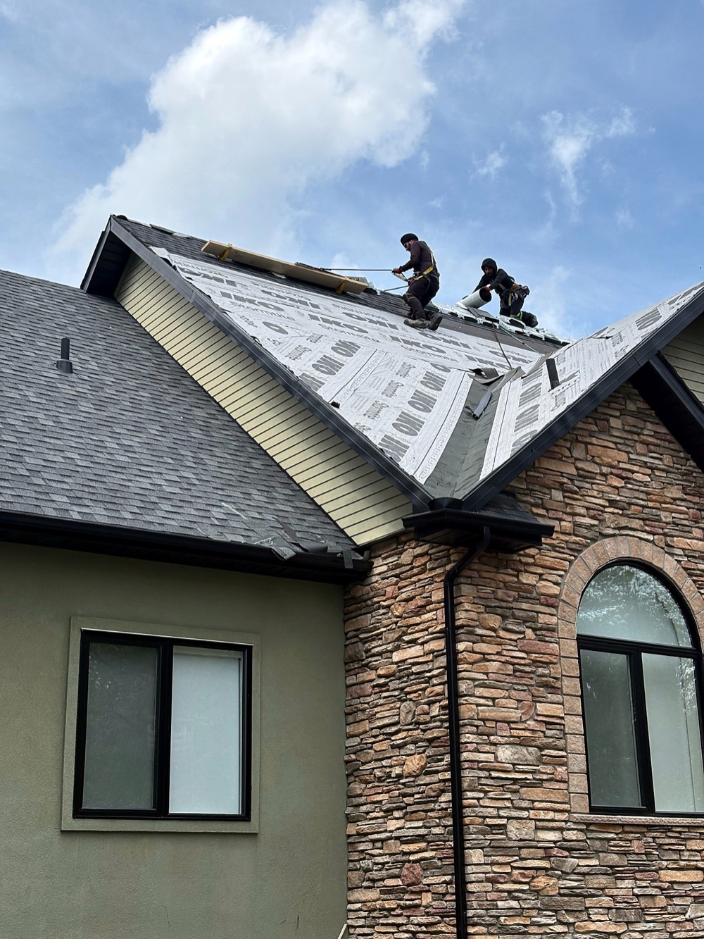 Two roofers on a steep gable laying underlayment on a stone-fronted house